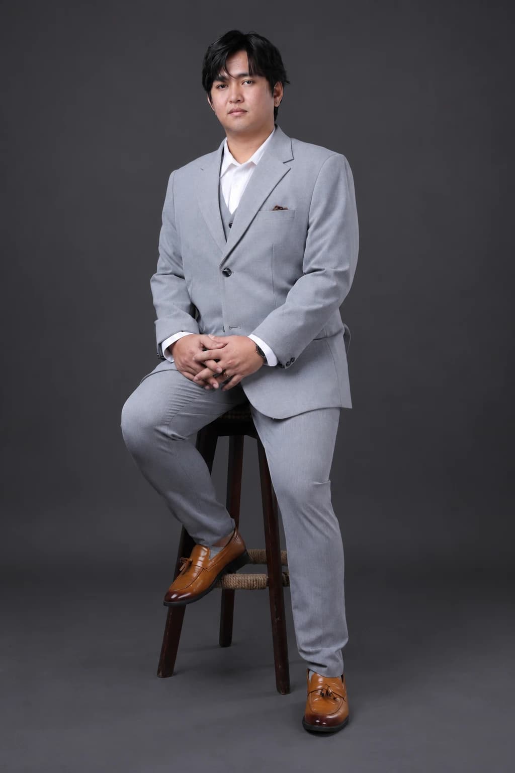 Edison seated on a tall stool in a grey suit, with hands clasped and brown shoes visible